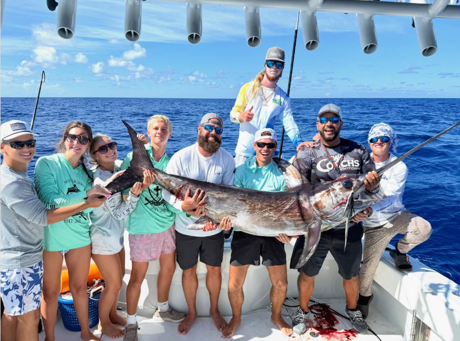 Group of people on a boat holding a large swordfish under a blue sky.