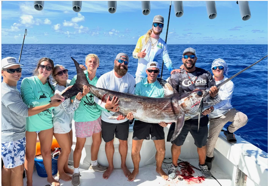 Group of nine people on a boat holding a large swordfish against a clear ocean backdrop.