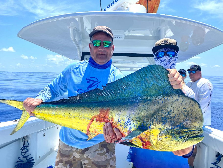 Two men on a boat holding a large, colorful fish against a blue sky and ocean backdrop.