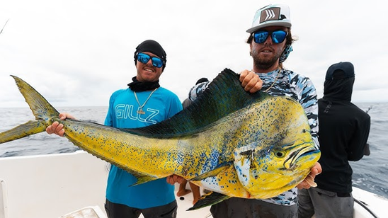 Two people on a boat holding a large, colorful bull dolphin fish against a cloudy sky.