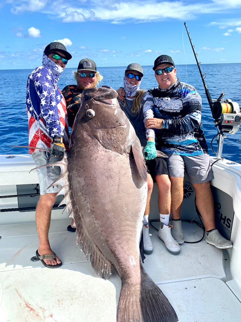 Four people on a boat holding a large fish with ocean in the background.