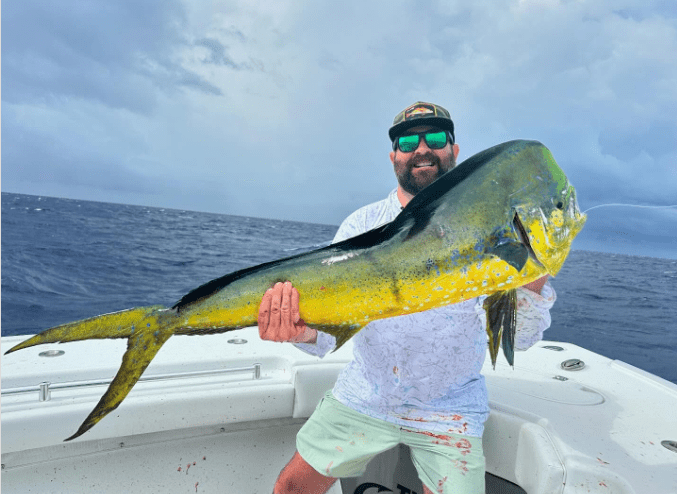 Man on a boat holding a large yellow and green fish against an overcast sky.