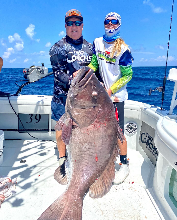 Two people on a boat holding a large fish with a blue ocean background.