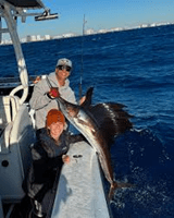 Two people on a boat holding a large sailfish against a backdrop of the ocean.