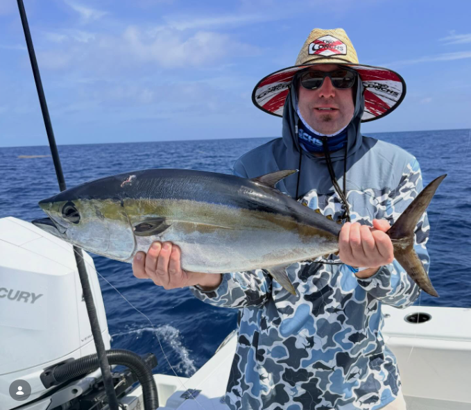 Person holding a blackfin tuna on a boat, wearing a camo shirt and wide-brimmed hat, with ocean in background.