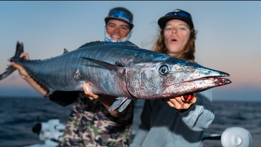 Two people holding a large fish on a boat at sunset.