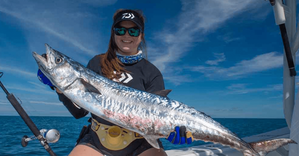 Person on boat holding a large fish, wearing a visor and sunglasses, blue sky in background.