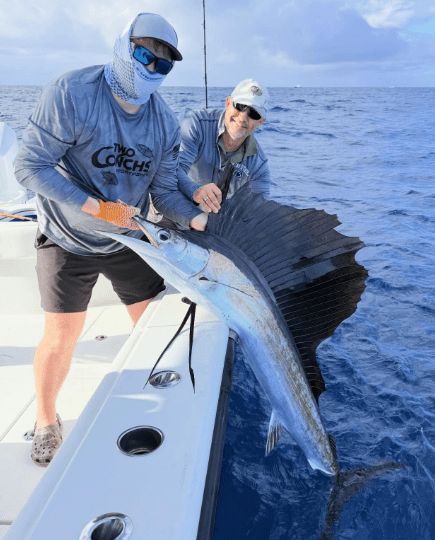 Two anglers on a boat hold a large sailfish over the water.