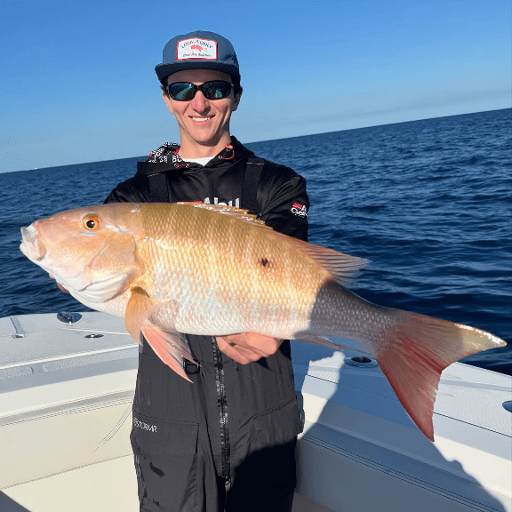 Person on a boat holding a large snapper fish against a clear blue sea backdrop.