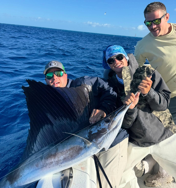 Three people on a boat holding a large sailfish in the ocean.