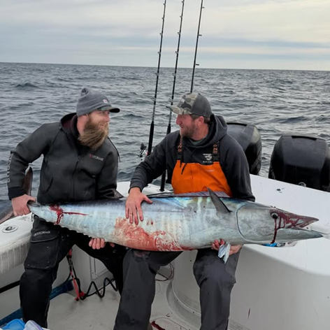 Two men on a boat holding a large fish with fishing rods in the background.