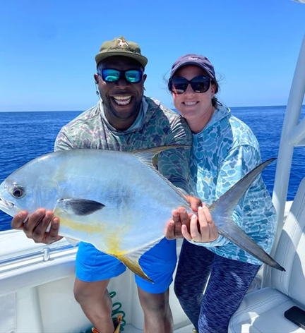 Two people smiling, holding a large fish on a boat under a clear blue sky.