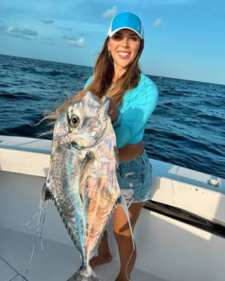 Person in blue cap and top holds a large African Pompano fish on a boat in the ocean.