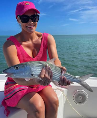 Woman on a boat holding a bonefish, wearing a pink outfit and sunglasses.