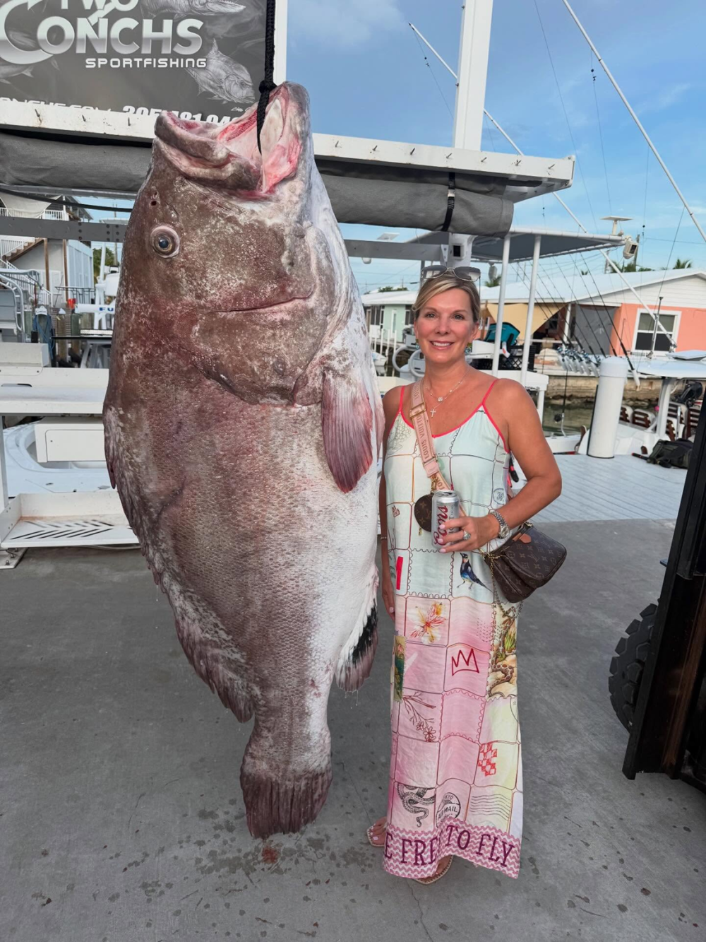 Woman in sundress stands beside large Grouper fish hanging from pier.