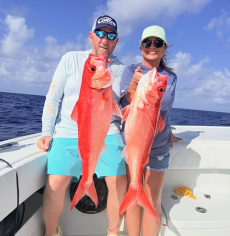 Two people on a boat holding large red fish, with blue sky and ocean in the background.