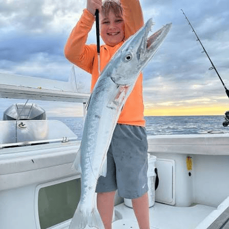 Child proudly holding a large barracuda fish on a boat at sea, under a cloudy sky.