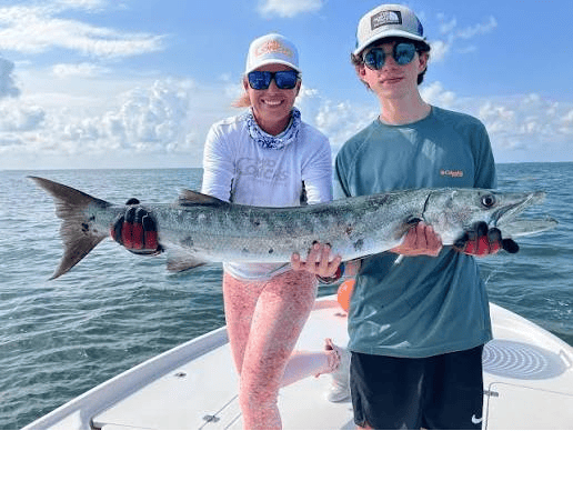 Two people on a boat holding a large barracuda fish together, smiling with the ocean in the background.