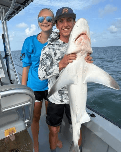 Two people on a boat holding a large shark on the ocean under a blue sky.