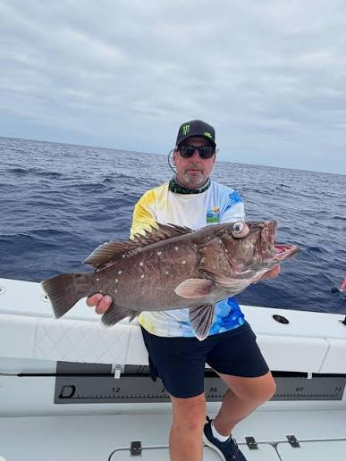 Person on a boat holding a large snowy grouper fish against a cloudy sky.