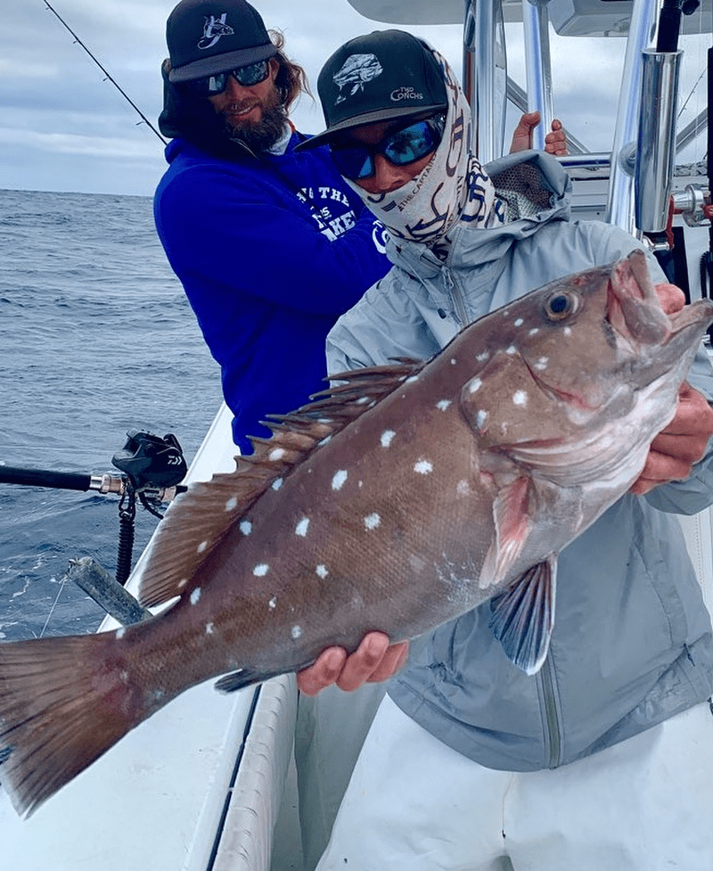 Two anglers on a boat, one holding a large snowy grouper fish with white spots.