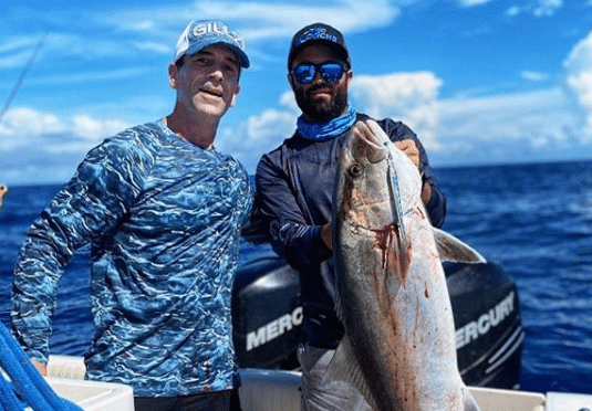 Two men on a boat holding a large amberjack fish with the ocean in the background.