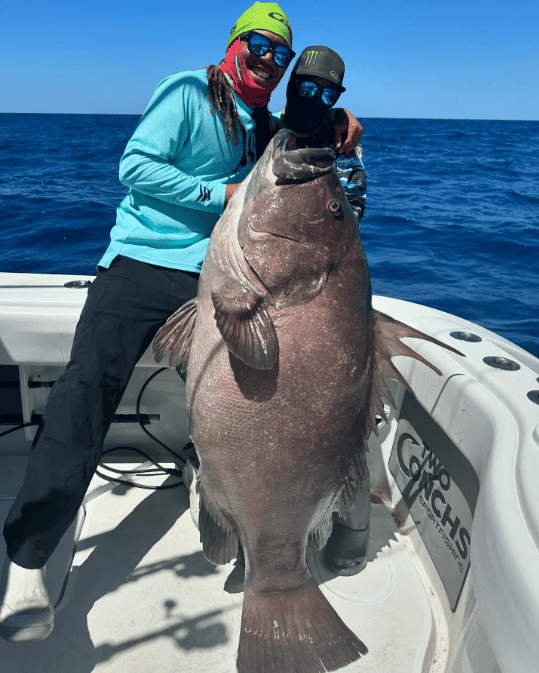Two anglers on a boat holding a Goliath Grouper fish against a blue sea backdrop.