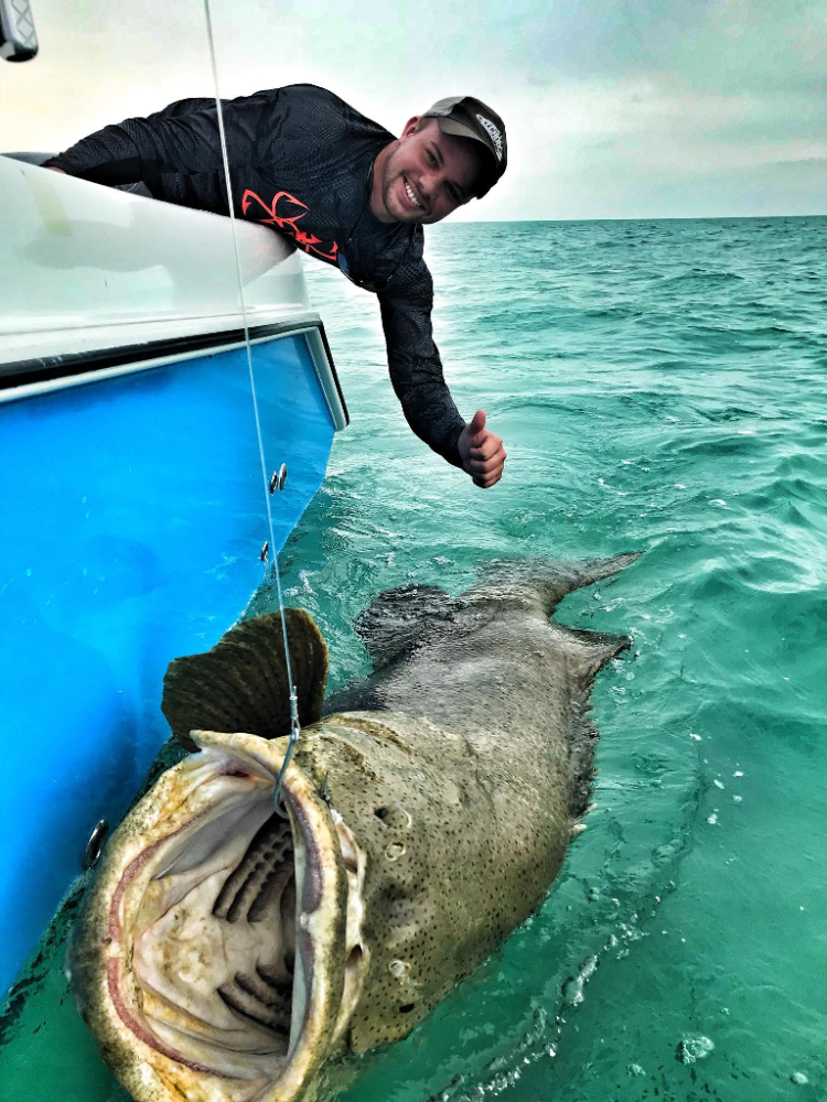 Person on boat gives thumbs up next to goliath grouper fish with open mouth in the ocean.